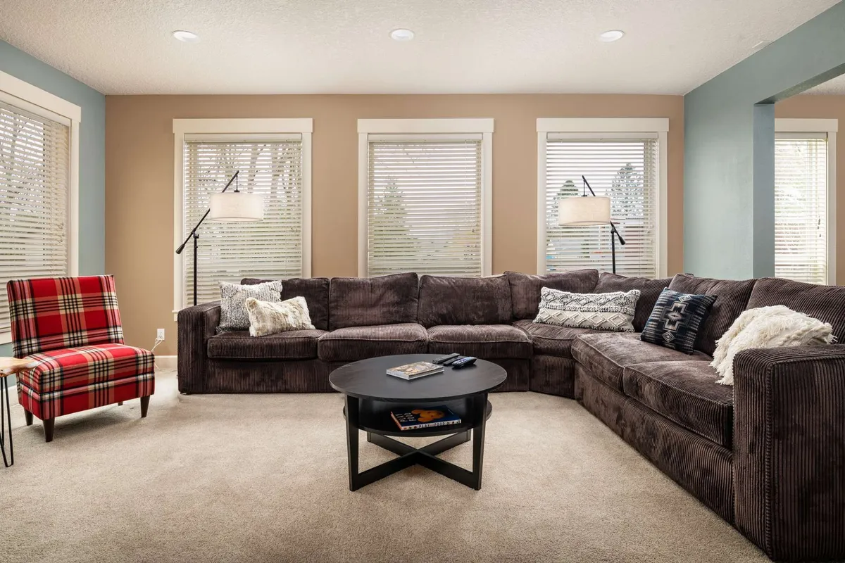 A spacious living area with a large sectional sofa, a plaid accent chair, and a coffee table, all bathed in natural light streaming through sizable windows. The space feels inviting and connects visually to the outdoors, suggesting a comfortable spot for guests (and their pets) to unwind after outdoor adventures.