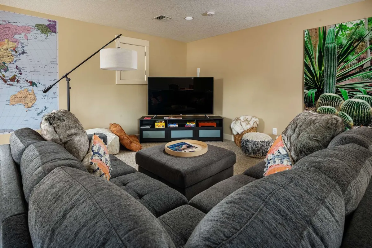 A spacious living area featuring a large sectional sofa, coffee table, television, and prominent world map artwork alongside plant-themed art. The room is bathed in natural light, creating a relaxed, inviting atmosphere ideal for unwinding after urban or outdoor activities.
