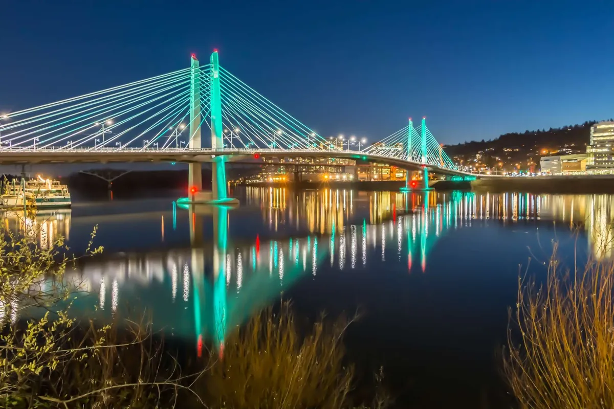 The scene shows an illuminated bridge reflecting on calm waters, surrounded by city lights. The riverside path and glowing backdrop evoke the ambiance of evening community events and leisurely strolls along the water.
