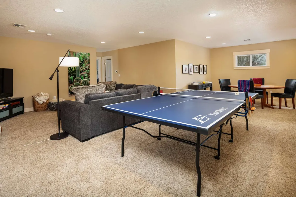 An open-concept recreation and living area featuring a ping pong table, cozy sofa, television, and a visible dining area in the background. The space is bright and arranged for both active play and relaxed socializing.