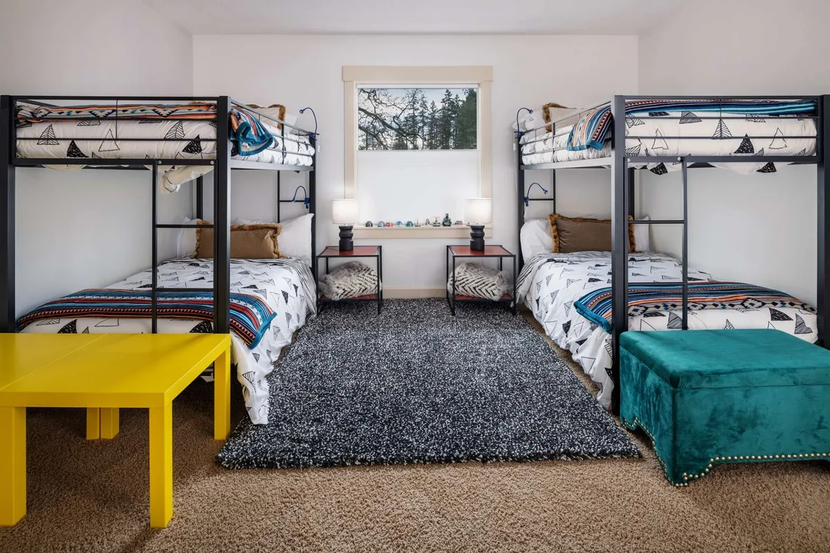 A cheerful bunk room featuring colorful bedding on built-in bunk beds, with a bright yellow table and a green ottoman providing space for play or reading. The room feels playful and inviting, ideal for children or groups of kids to enjoy sleepovers and creative adventures.