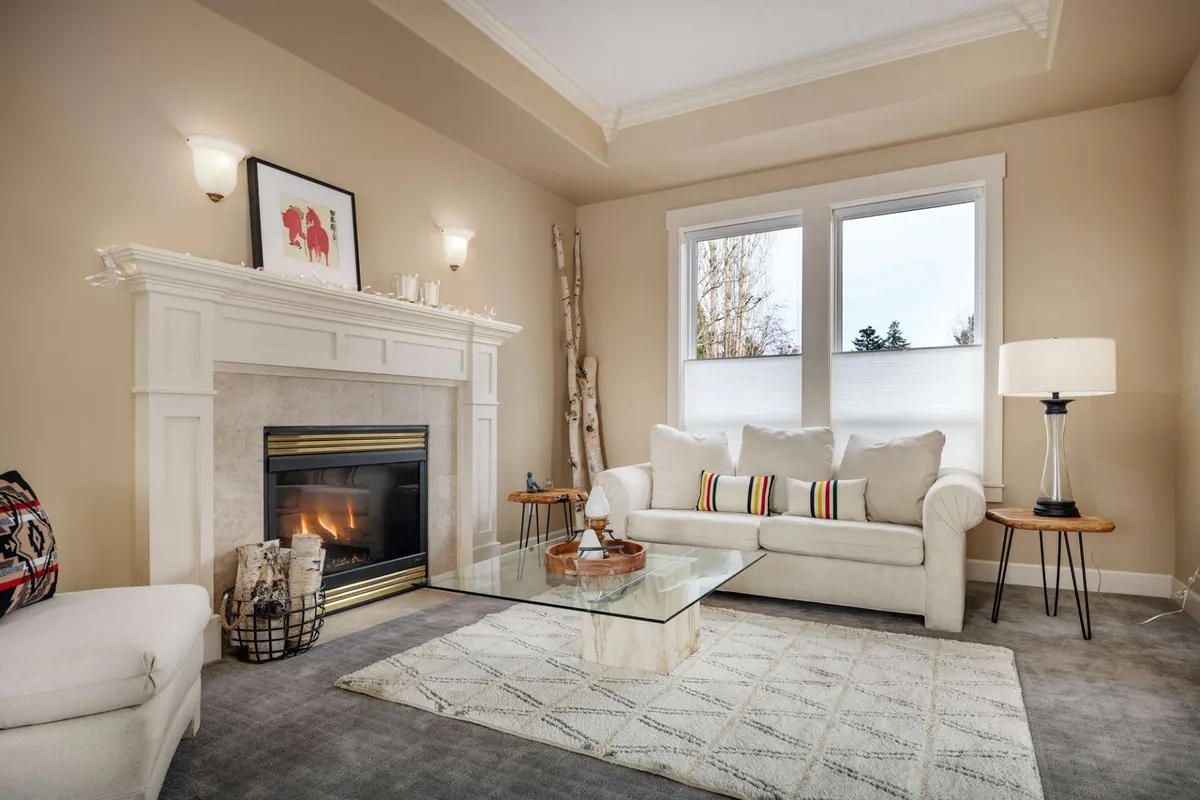 A cozy living space anchored by a fireplace, with a comfortable sofa, a coffee table, vibrant artwork, and sunlight streaming through generous windows. The arrangement encourages relaxation and conversation.