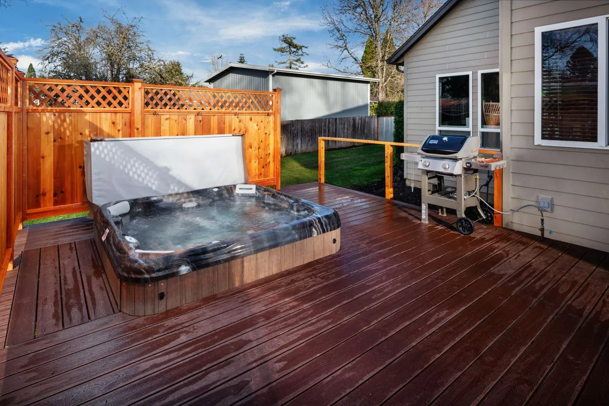 This exterior shot showcases a wooden deck with a hot tub and grill set against the backdrop of a tranquil sunset. The deck is designed for relaxing and socializing, with the hot tub inviting guests to soak under the open sky, while the grill hints at alfresco dining and lively gatherings.