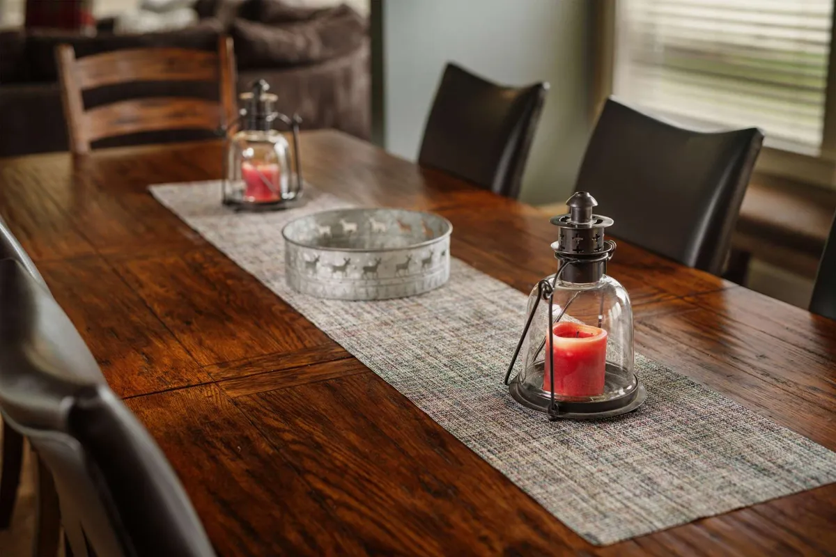 A warmly-lit dining area featuring a large wooden table set for a meal, surrounded by comfortable seating. Lanterns with candles and a decorative bowl serve as the centerpiece, while subtle décor hints at a cozy, inviting space ideal for shared meals or celebrations.